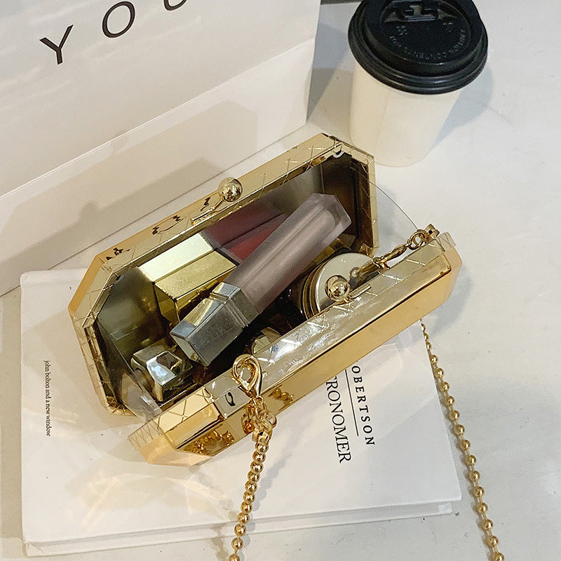 The Baazuu Golden Phantom box clutch in gold. This modern, octagonal evening bag features a minimalist, geometric design in a hard-shell metallic finish, complete with a chain strap.