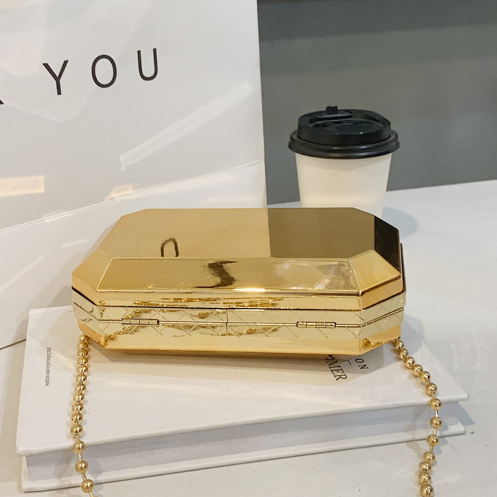 The Baazuu Golden Phantom box clutch in gold. This modern, octagonal evening bag features a minimalist, geometric design in a hard-shell metallic finish, complete with a chain strap.
