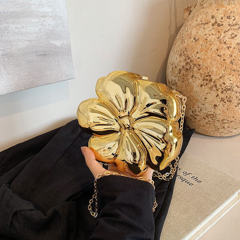 The Baazuu Bloom Bling floral clutch in a metallic gold finish. This sculptural, flower-shaped handbag features a hard-shell design and a chain strap, perfect as a modern statement piece.