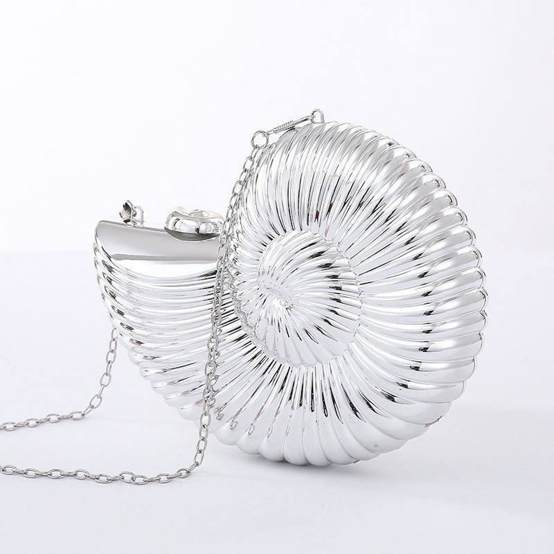 The Baazuu Auréelle conch bag. This unique, shell-shaped clutch features a sculptural, spiral design in a metallic silver finish with a chain strap, perfect as an artistic statement piece.