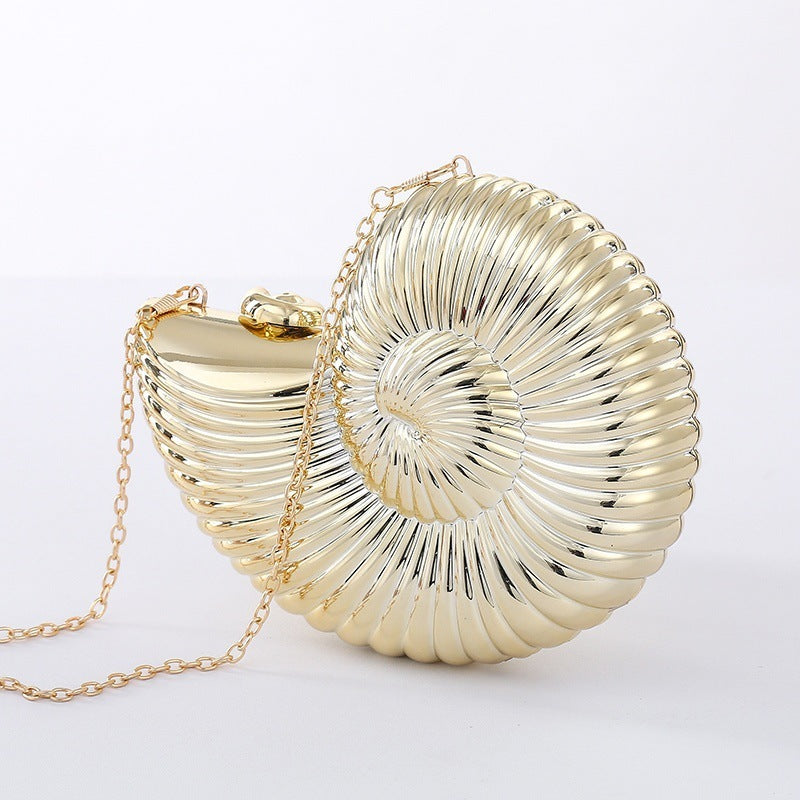 The Baazuu Auréelle conch bag. This unique, shell-shaped clutch features a sculptural, spiral design in a metallic gold finish with a chain strap, perfect as an artistic statement piece.