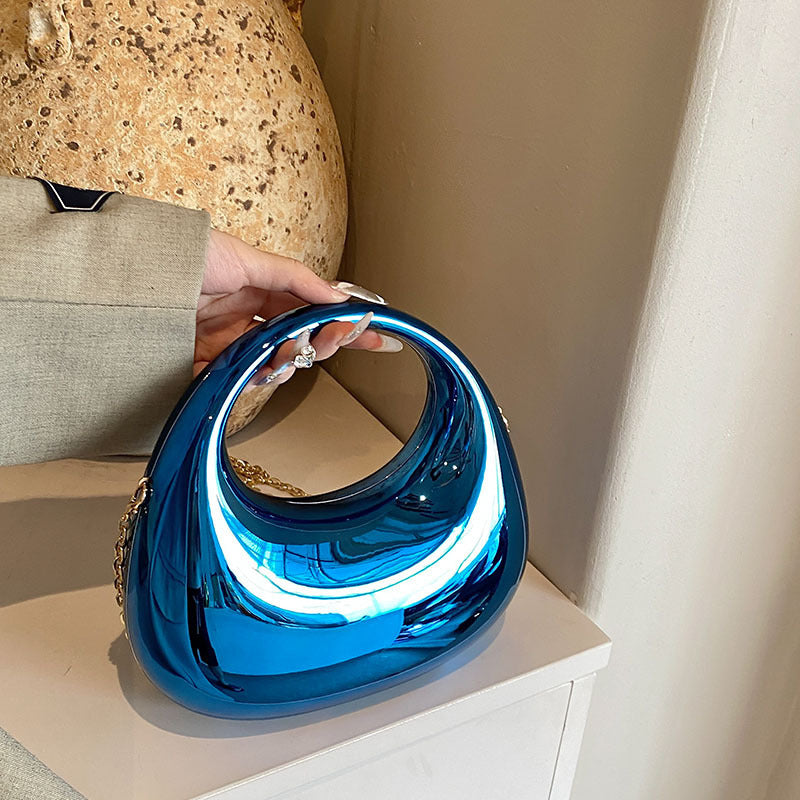 Front view of the Golden Eclipse handbag in a metallic blue finish. The bag has a unique crescent shape with a large circular handle cutout and a delicate gold chain strap. brand baazuu