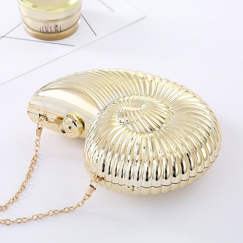 The Baazuu Auréelle conch bag. This unique, shell-shaped clutch features a sculptural, spiral design in a metallic gold finish with a chain strap, perfect as an artistic statement piece.